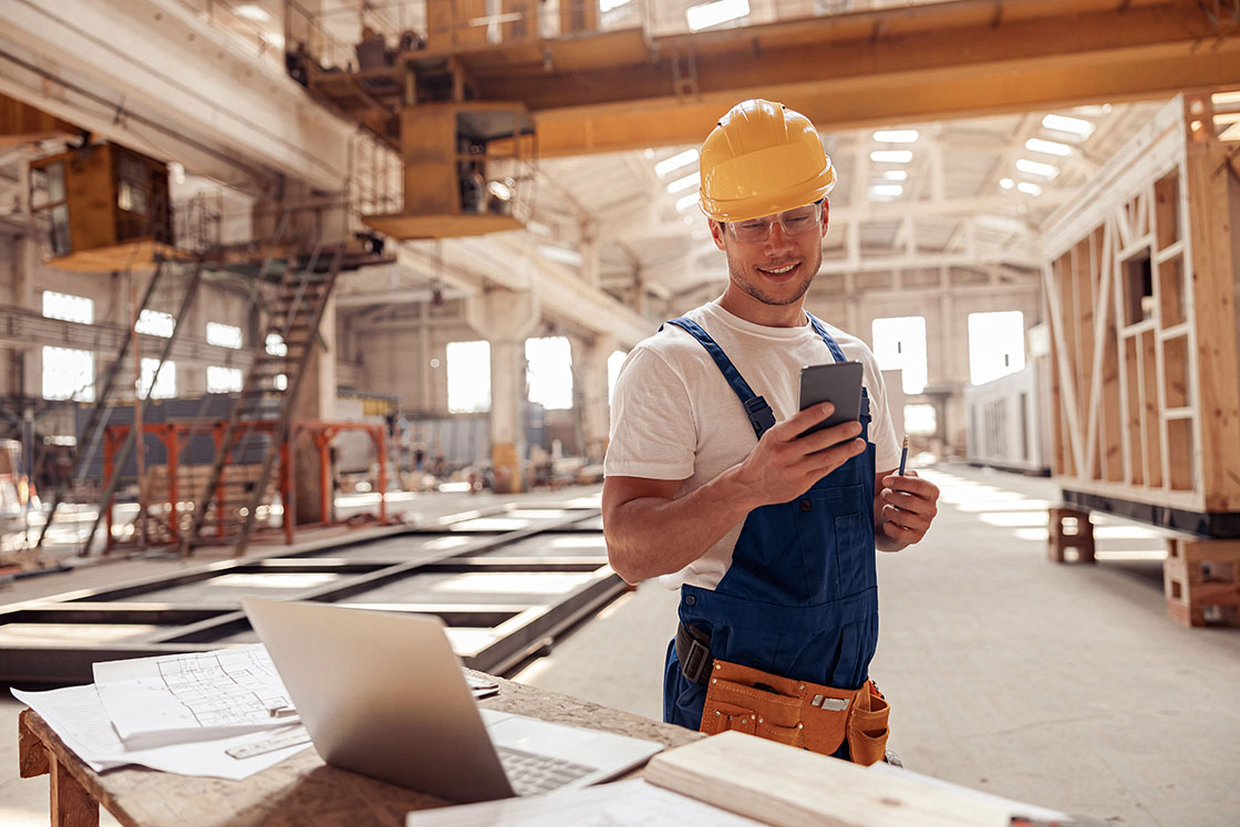 A construction worker holding a cell phone in a warehouse.